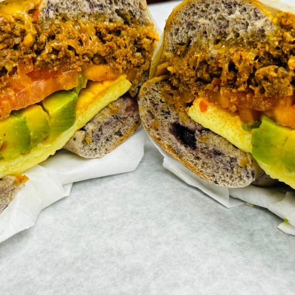 Close-up of a New York Bagel Cafe bagel sandwich, cut in half, revealing avocado, egg, and meat.