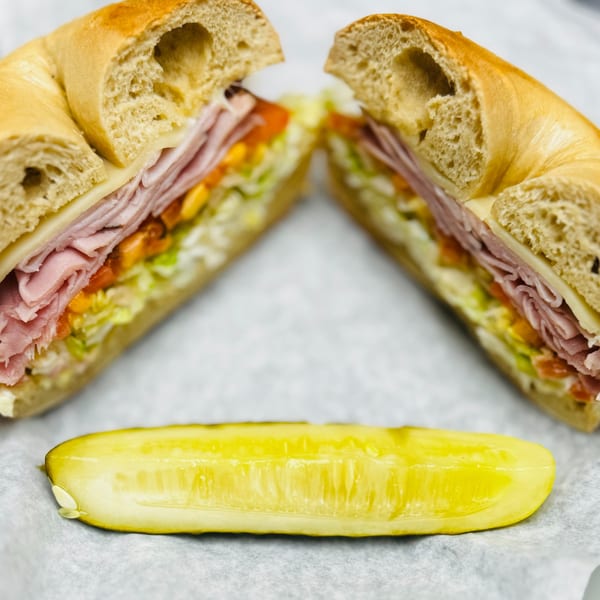 New York Bagel Cafe's bagel sandwich, cut in half, with ham, cheese, lettuce, tomato, and a pickle.