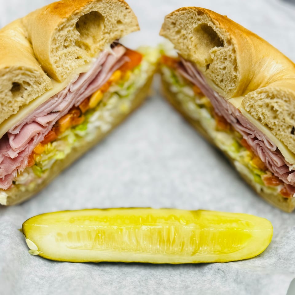 New York Bagel Cafe's bagel sandwich, cut in half, with ham, cheese, lettuce, tomato, and a pickle.